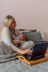 Young woman in comfortable home clothes works on laptop with a baby in her arms in bedroom.