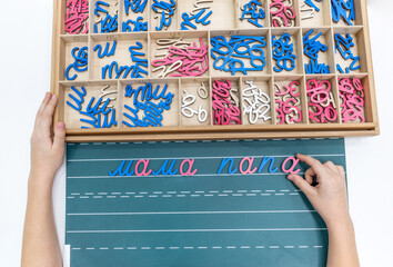 Top view of kids hands building words by using colored Montessori movable alphabet from the wooden tray on blackboard.
