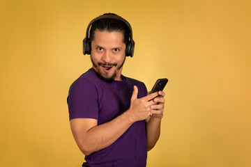 Portrait of man with funny face using headphone with yellow background. Man using smartphone