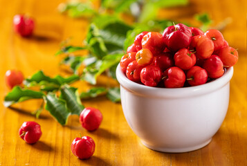 Acerolas maduras dentro de um pote de cerâmica branco com um galho da aceroleira desfocado no fundo.