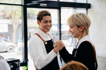 Cheerful young Asian man and woman hairdresser wearing apron uniform in modern salon giving high-five to each other on celebrating success of salon