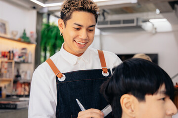 Happy young Asian male hairdresser smiling wearing apron in modern salon holding brush and scissors...