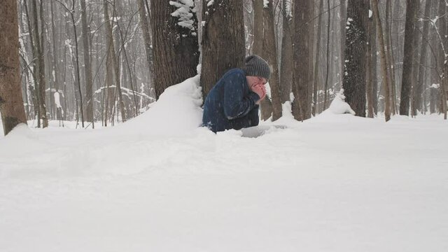 It is snowing heavily. Drunk caucasian man without jacket freezes in forest sitting on snow near tree. Concept of problems of homeless people and people who have harmful addictions to alcohol, drugs