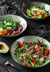 Fresh salads with avocado, shrimps, beef steak, tomatoes, green leaves, arugula, spinach, chicken breast and sweet sauce on dark wooden background. Healthy food, clean eating, closeup