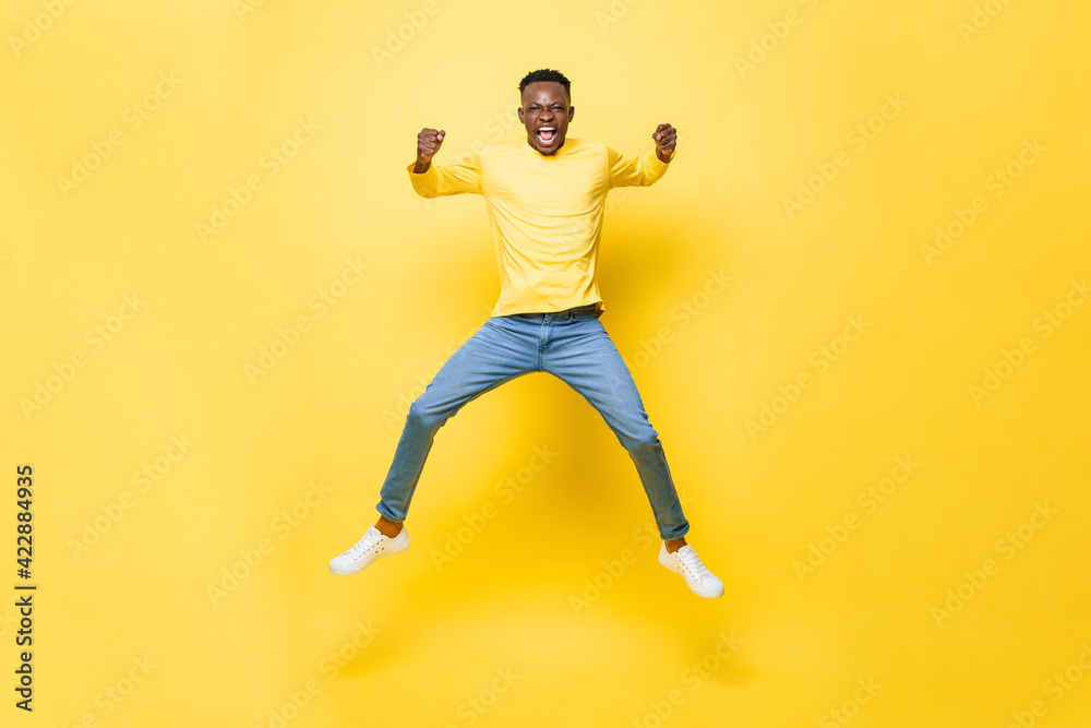 Wall mural Young cheerful excited African man jumping and screaming with clenching hands on isolated yellow studio background