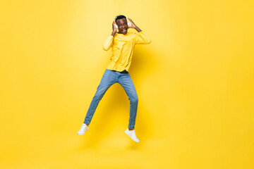 Happy young African man wearing headphones listening to music and jumping in yellow isolated studio background