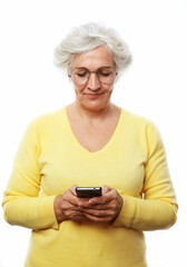 Fototapeta premium Lovely senior lady in casual outfit cheerfully smiling and looking at camera while standing on white background and using modern smartphone