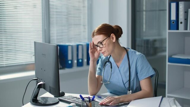 headache and eye strain at work, female doctor in blue uniform works at computer then takes off glasses and massages irritated eyes