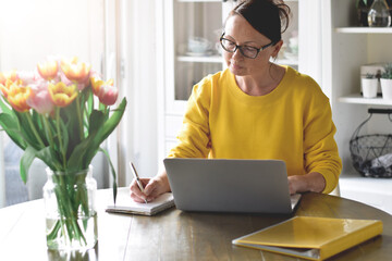 Woman in yellow sweater sitting at a desk writing notes while working on laptop computer