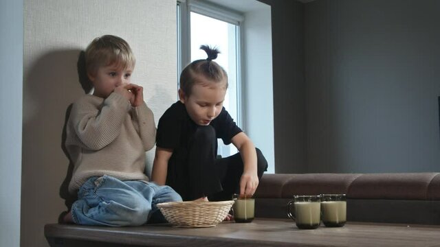 Two little boys sit on table eat a cookies with milk
