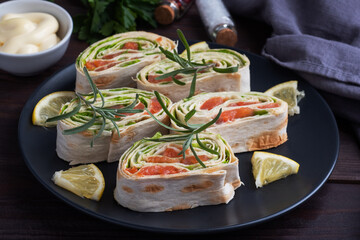 Rolls of thin pita bread and red salted salmon with lettuce leaves on a black ceramic plate, dark wooden background.