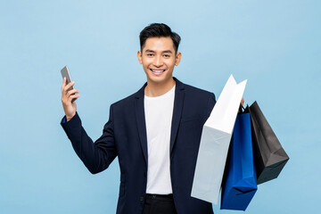 Young handsome southeast Asian man holding mobile phone and shopping bags in light blue studio...
