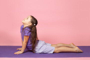 Obraz na płótnie Canvas full-length portrait of a girl in profile doing yoga and bhujangasana or upward dog pose, Cobra Pose on a purple mat isolated on a pink background.