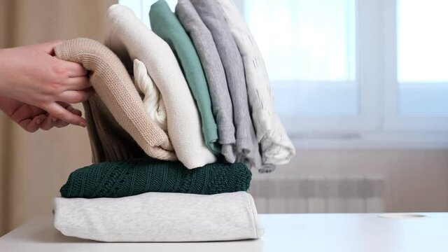 Woman takes sweater from middle of folded knitwear clothes and rest stack falls on white table against large window in brightly lit room extreme closeup