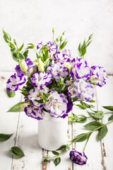 A bouquet of eustoma flowers in a white vase on a white wooden table.