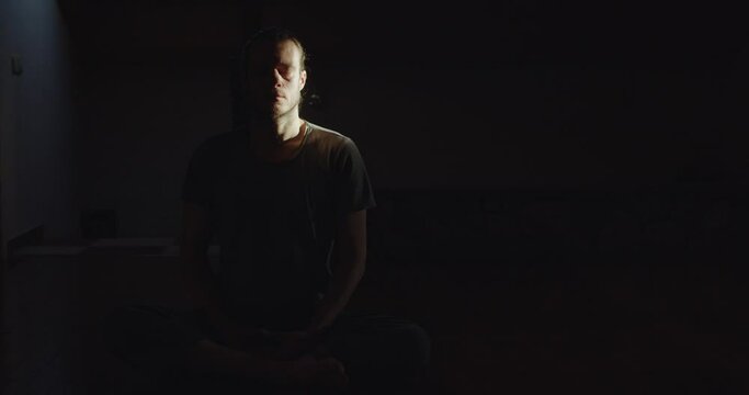Portrait of man sitting in lotus asana meditating alone in dark room black background copy space. Pensive calm yogi practicing yoga meditation indoor morning light text. Tranquility calmness wellness 
