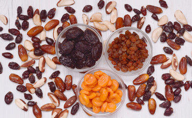 Prunes, dried apricots and raisins in glass plates, which are on the table, and nuts, dates and rose hips are scattered around