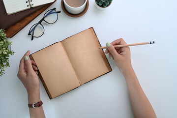 Overhead shot of young woman holding pen writing on empty notebook at white office desk.