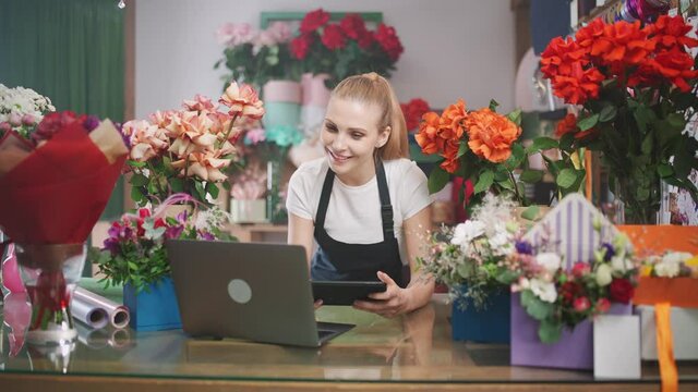 Flower delivery service, female florist discusses the order by talking on video call, using a laptop and screen tablet in a flower shop, working online, the female chooses flowers for the customer.