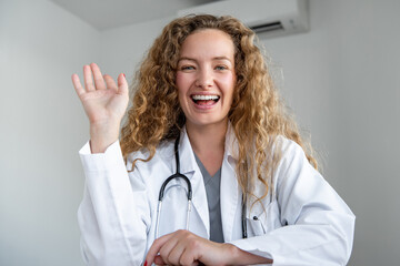 Happy smiling young female doctor waving hand greeting patient online via video call, home medical...