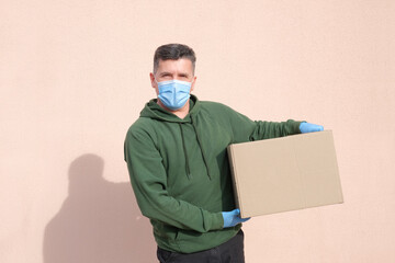 Middle-aged delivery man in medical mask and gloves holding and carrying a cardbox on pink wall background. Volunteer delivery of food or medicine during the coronavirus pandemic