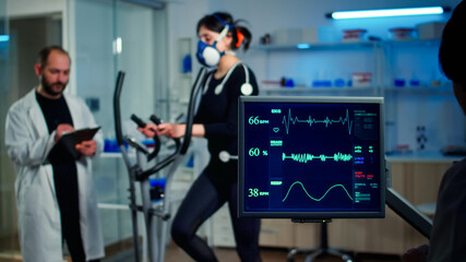 Specialist sport researcher monitoring heart rate on athlete while woman with mask running on cross trainer talking with medical doctor. Physician using tablet controling EKG data in modern laboratory