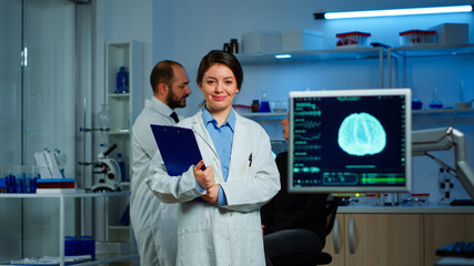 Portrait of scientist neurologist researcher looking at camera smiling while coworker discussing with patient in background about brain functions, nervous system, tomography scan working in laboratory