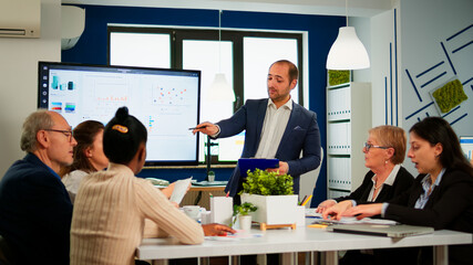 Manager pointing on digital interactive whiteboard with growth analysis, charts, statistics and data. Diverse people working in broadroom during conference meeting discussing company problems.