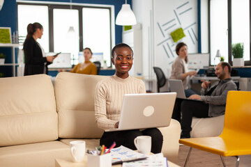 African woman manager sitting on couch in front of camera smiling while team working in background with financial reports. Diverse coworkers talking about startup financial company in modern office.