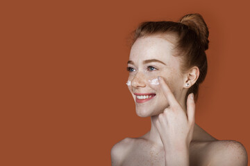 Caucasian woman with red hair and freckles applying anti aging cream on her cheeks smiling on a red wall with free space