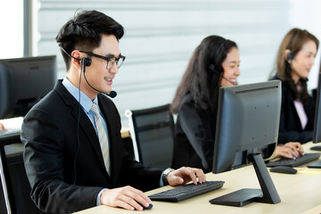 Cheerful Asian man in headset smile with cheerful and happy while working on computer at desk with female coworkers in help desk office. Sevice mind and ready  for marketing support concept