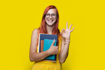 Ginger freckled student is gesturing the okay sign on a yellow studio wall holding books and wearing a dress and glasses