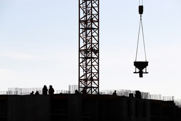 Silhouettes of workers and tower crane with cargo on construction site against the sky. Housing construction, builders working on scaffolding