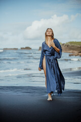 Fototapeta na wymiar Photoshoot of a blonde girl in a blue dress on the Bali beach with black sand