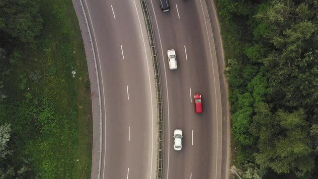 Flying Over A Curved Motorway With Traffic: White And Black Cars And Trucks Move Along The Lanes Of The Highway Among The Green Foliage - Aerial Overhead Shot. Traffic On Country Road On A Summer Day.