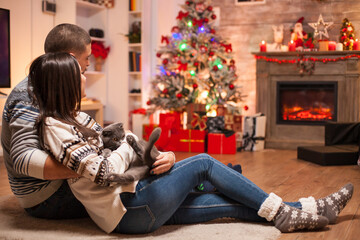 Obraz na płótnie Canvas Couple in matching clothes and their scottish fold sitting together on the floor in front of fireplace celebrating christmas.