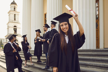 Smiling young graduated woman wearing gown and cap standing with diploma scroll in raised hand on background of group of students