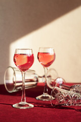 two glasses of red alcoholic drink among empty glasses lying on the table, light and shadow on the background, berry liqueur or cocktail among fallen tableware, copy space, minimalism