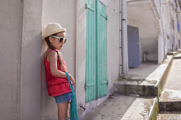 Stylish girl of gen Z in summer hat and mint sunglasses with mesh bag standing near the wooden door at the street of small old town, France, summer family trip, extending vacation with local immersing