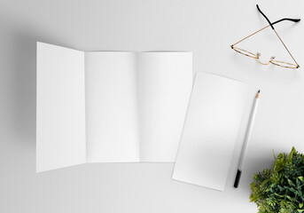 Plain white trifold brochure on an isolated background surrounded by white pencil glasses and flower pot
