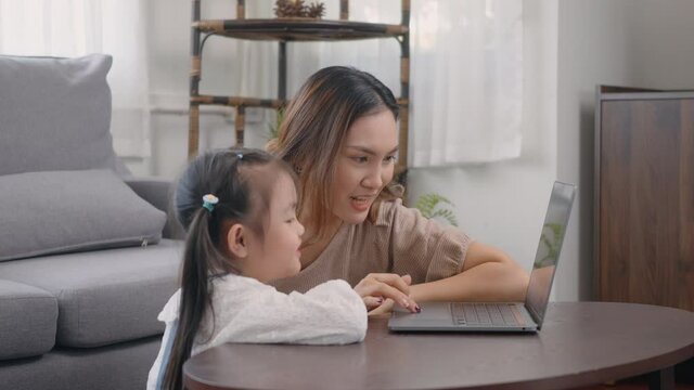 Asian young mother with laptop computer teaching her kid to learn or study online in living room at home, Mum and little preschool daughter learning online on computer, family homeschooling online