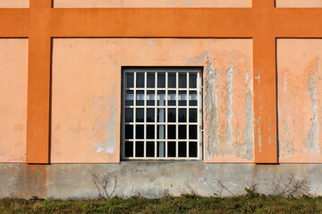 New window with white plastic frame and metal bars protection mounted on colorful old dilapidated industrial factory building wall with cracked facade and uncut grass in front