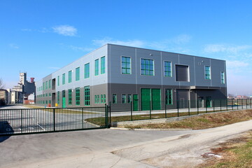 Fototapeta na wymiar Newly built modern grey and green production industrial building surrounded with closed metal fence and large old concrete storage silo building in background on warm sunny winter day