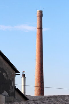 Red bricks tall industrial chimney with metal stairs mounted on side and three lightning rods on top rising high above old abandoned factory building with cracked facade at local industrial complex