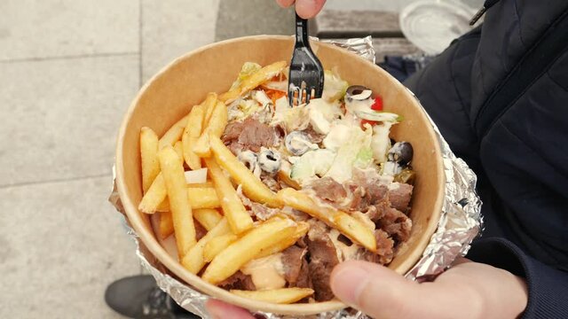 Man on a street sit eating takeaway food box with kebab meat with salad and fried potatoe chips from disposable tableware