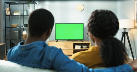 Close up of wife and husband sitting at home together watching TV with chroma key. Back view of African American couple man and woman sitting on sofa watch television program, green screen