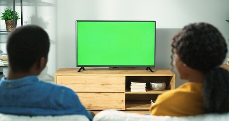 Back view of young African American family couple watching TV with chroma key while resting at home sitting on sofa in room. Wife and husband spending time together watch television with green screen