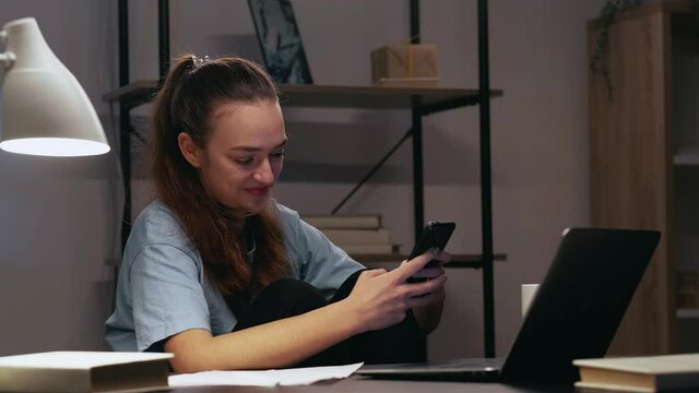 Mobile chat. Distance work. Job procrastination. Online communication. Relaxed lazy female employee using phone texting message smiling killing time at evening workplace with laptop in home office.