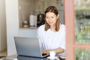 Happy young woman. remote work on a laptop computer from your favorite kitchen at home. White t-shirt and blue jeans, brunette woman. the online courses teacher prints a text message to their students