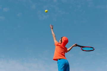 Child boy tennis player in orange sportswear with racket learning to performs serve ball. Kids sport tennis game, training at school or club. Child athlete in action. Background copy space